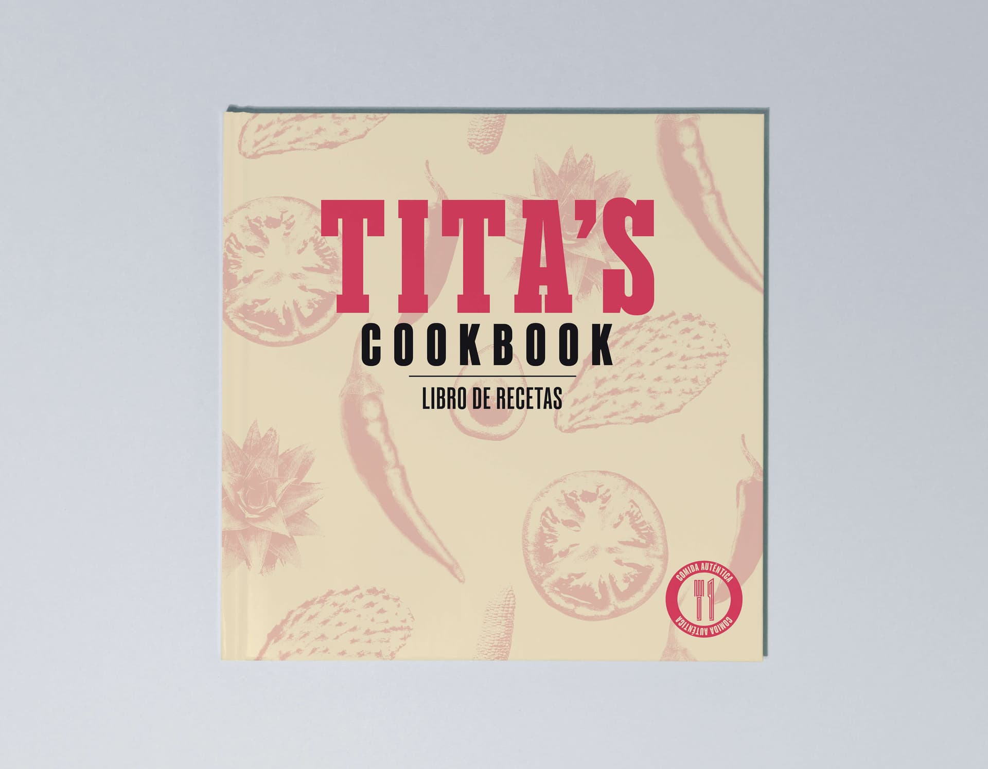 Hardcover mockup of Tita's Cookbook, cream cover with pink and black "Tita's Cookbook / Libro de Recetas" lettering over an illustrated pattern of chiles, tomatoes, and nopales, with a red "comida auténtica" seal in the corner